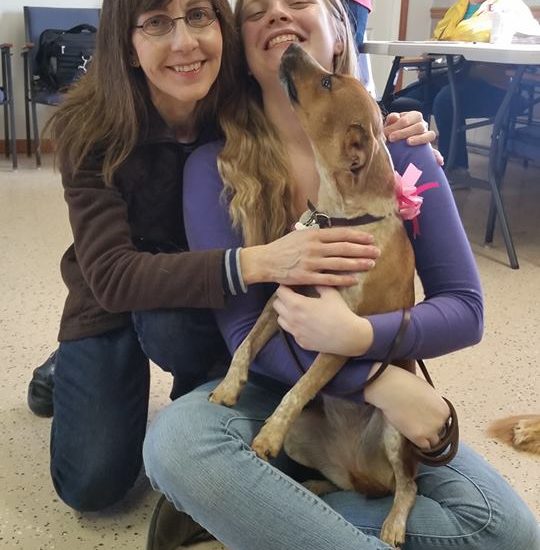 Two women hugging a dog in a room.