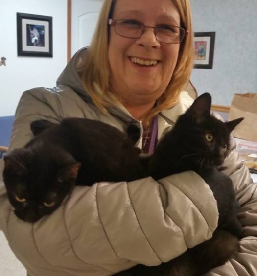 A woman holding two black cats in her arms.