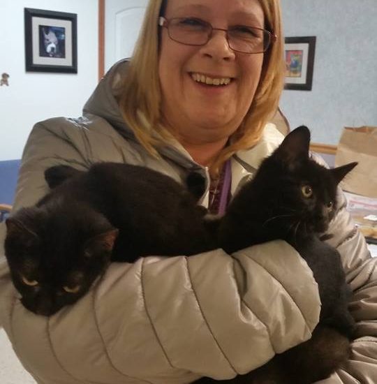 A woman holding two black cats in her arms.