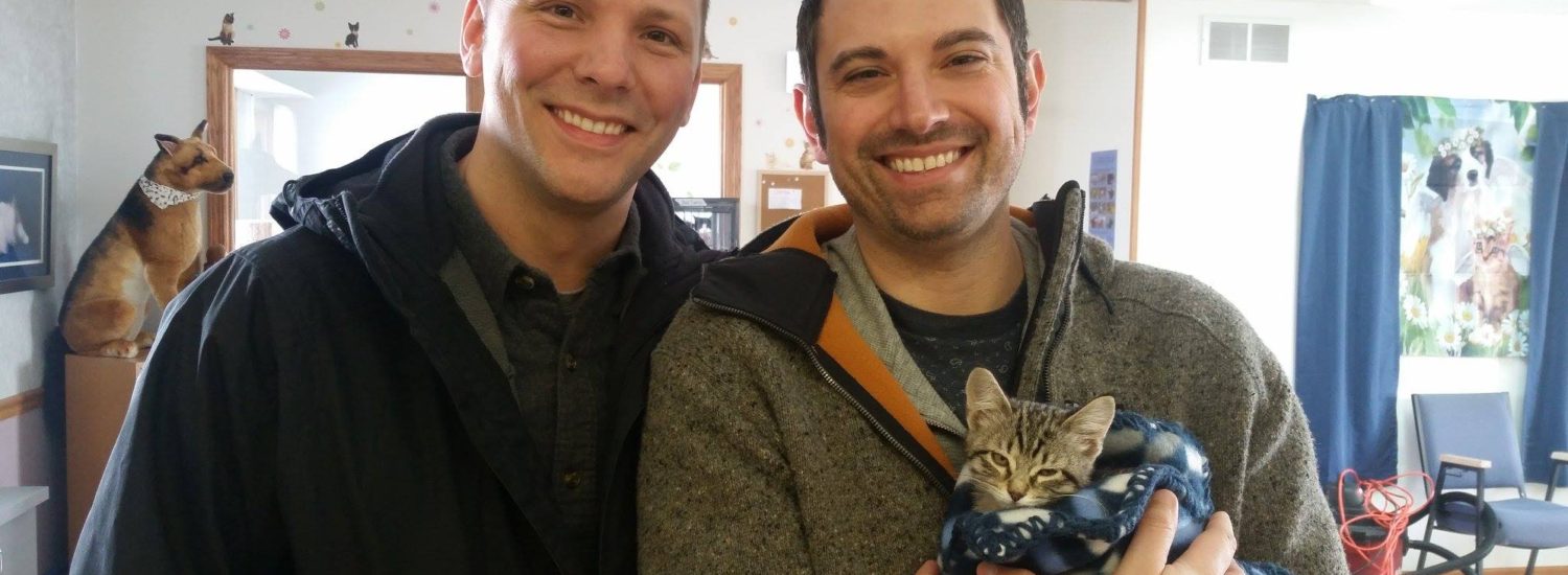 Two men smiling while holding a kitten in their arms.