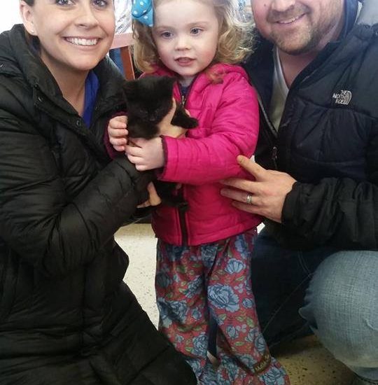 A man and woman are posing with a little girl and a black cat.