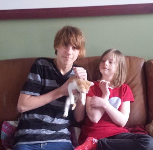 A boy and girl sitting on a couch holding a cat.