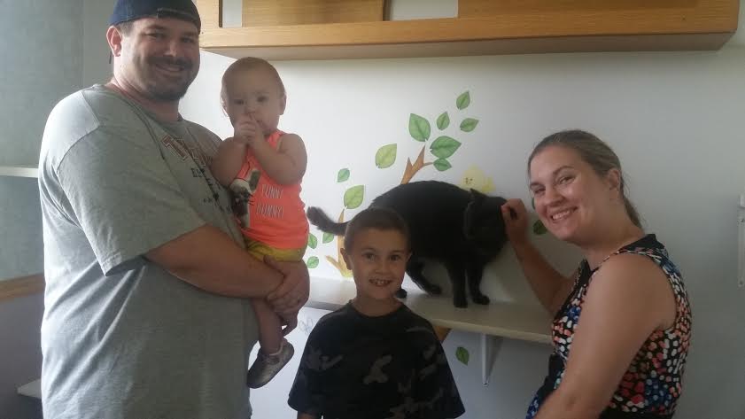 A man and woman pose with a cat in a kitchen.